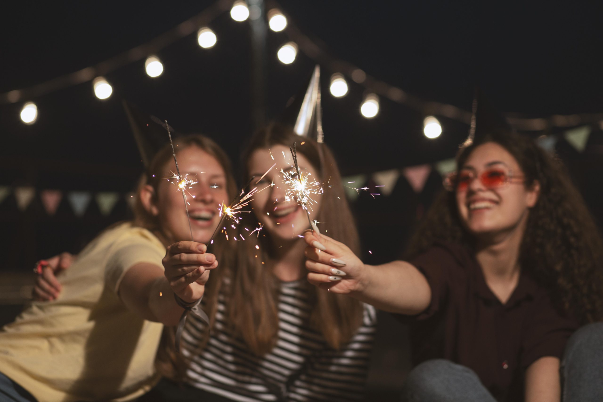 Três garotas comemoram o ano novo com velas. Ao fundo está de noite com algumas lâmpadas dispostas em um varal e iluminando o local.