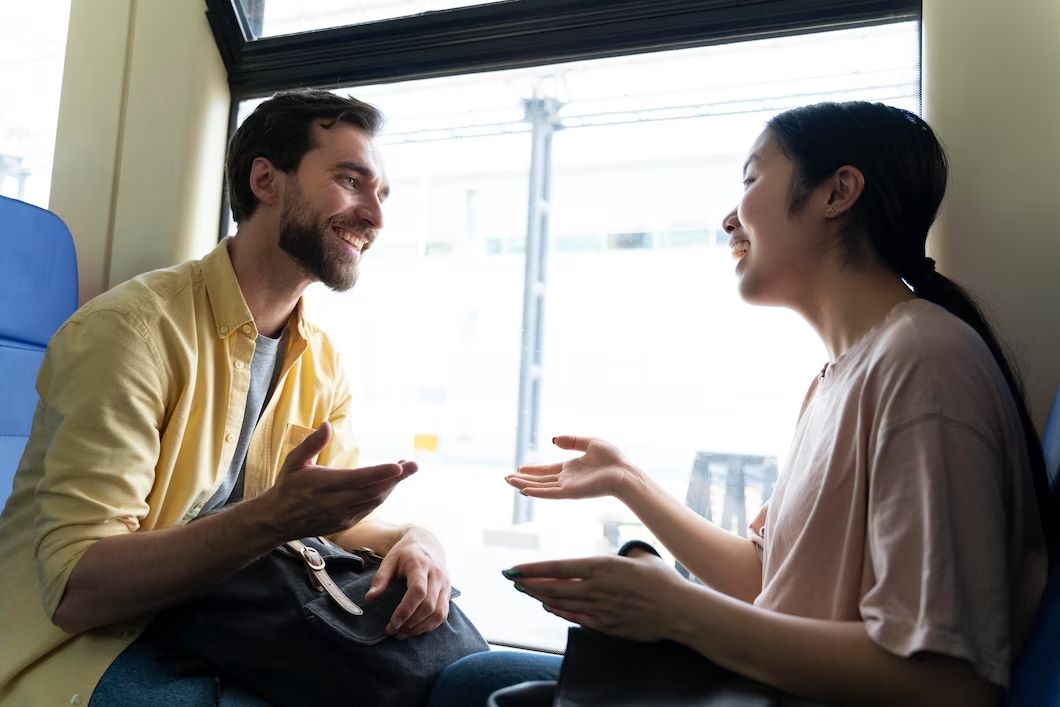 Dois jovens viajando e conversando em inglês dentro de um trem.