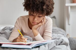 Uma jovem com cabelos crespos concentrada, escrevendo em um diário ou caderno enquanto está sentada na cama, refletindo sobre seus diários de aprendizado.