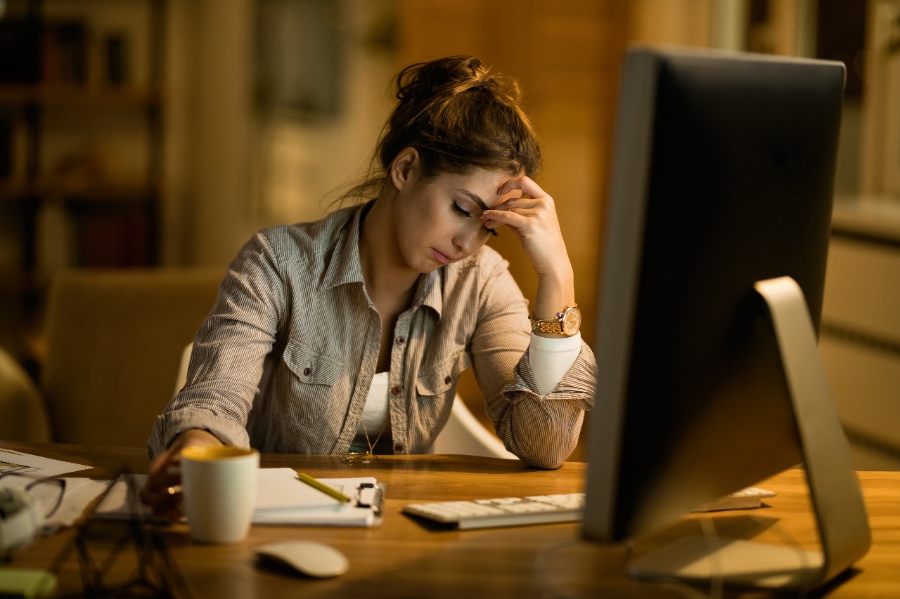 Uma mulher sentada à mesa de trabalho à noite, parecendo exausta e com a mão na testa, ilustrando o sentimento de burnout.